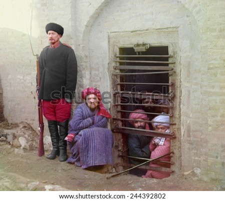 Traditional Central Asian prison, with cells cut into the ground. Inmates looking out through bars, guard stands by with Russian rifle, uniform, and boots. ca. 1910. photo by Sergei Prokudin-Gorskii,