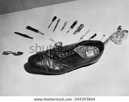 Law enforcement photo of a shoe with metal box in cut out section of heel used to smuggle drugs into a detention facility. For dramatic effect the shoe is surrounded by drug paraphernalia. 1955.