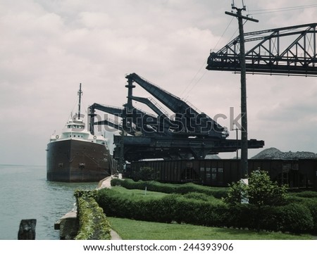 Great Lakes freighter unloading iron ore at the Pennsylvania Railroad ore docks in Cleveland Ohio. One \'Hulett\' unloader is lowering its bucket into ship\'s hold, another loads a railroad car. 1943.