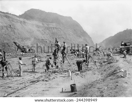 Panama Canal construction showing workers drilling holes for dynamite in bedrock, as they cut through mountains of the Isthmus. Steam shovels in the background move the rubble to railroads car. 1913.