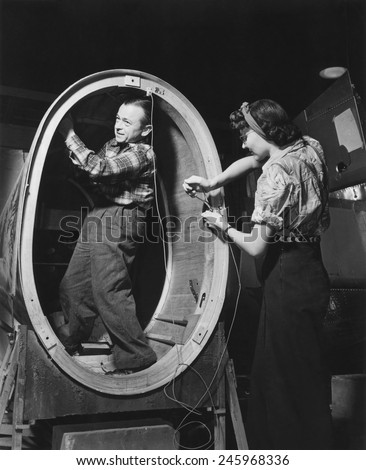A small man installs control wires inside the fuselage of a \'Valiant\' basic trainer while a woman works standing outside. Vultee Aircraft Downey Plant California. Ca. 1942-43.