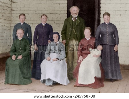 A Latter Day Saint plural marriage portrait. A middle aged man poses with his elderly mother, and five wives of various ages, one holding a baby. Photo by John P. Soule, ca. 1885 with digital color.