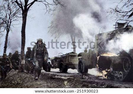 German soldiers passing burning U.S. equipment in the beginning of the Battle of the Bulge. Dec. 16-22, 1944. Still from a captured German film with color added later. B&W Photo with oil color.