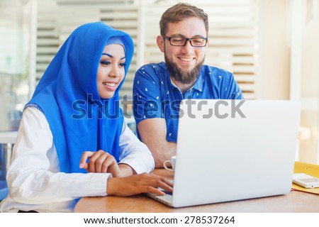 asian muslim woman and caucasian man looking at screen of laptop and working together