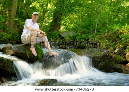 Happy young man sitting at a beautiful creek and cooling his foot in the water