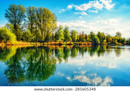 Tranquil landscape at a lake, with the vibrant sky, white clouds and the trees reflected symmetrically in the clean blue water