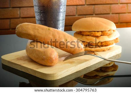 pork sausage, chicken hamburger and glass of cola on wooden cutting board with Knife and fork on top table, reflections