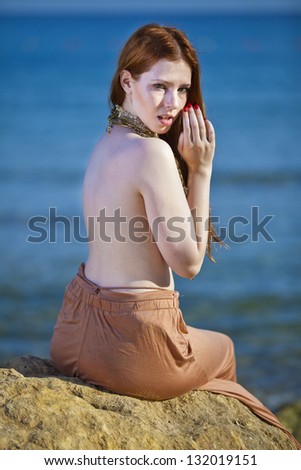 A beautiful woman with pale white skin and long red hair sitting on a rock semi-nude at the beach