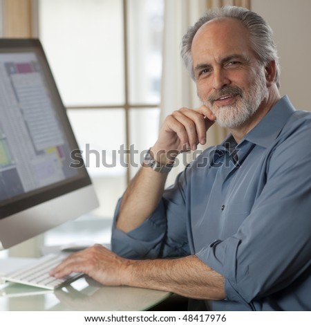 Portrait of a businessman dressed in casual clothing and sitting in front of a computer. He is smiling at the camera with one hand on the keyboard and the other near his face. Square format.