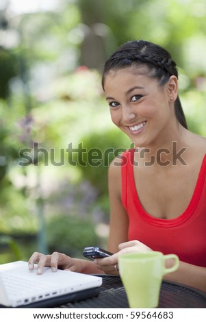 Young Asian woman sits at an outdoor mesh table with a laptop, mobile phone and coffee cup. She is smiling at the camera. Vertical shot.