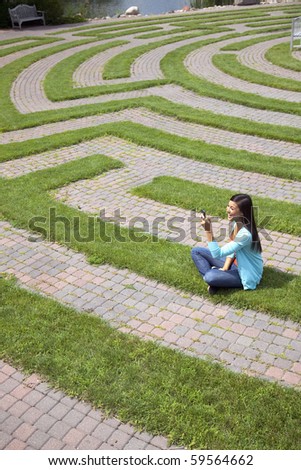 Beautiful young Asian woman laughs at a text message on her cellphone while sitting in a grass labyrinth.
