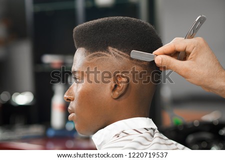 Side view of serious man with stylish modern haircut looking forward in barber shop. Hand of barber keeping straight razor and cutting trendy stripes on head of client. Concept of shaving.