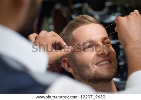Closeup of process of threading procedure in barber shop. Professional barber correcting shape of brows with threads to smiling young man sitting in chair. Concept of eyebrows care.