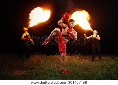 sportsman is training on the background of the fiery show, stand on one leg, hand raised over head, boxer in red shorts and red boxing gloves, muscular body, the night training, workout on open space
