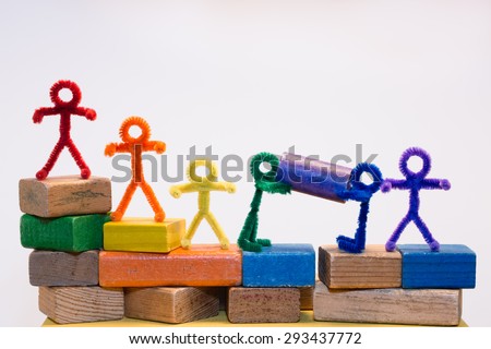 A row of rainbow pipe-cleaner people across children\'s building blocks with two helping each other to move a block. Wonderful photo for multicultural, international ideas and concepts.