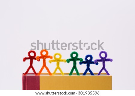 A row of rainbow colored pipe-cleaner people on top a text book. They seem to be holding hands as if helping one another. A great photo for multicultural and international ideas and concepts.