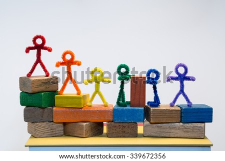 A row of rainbow pipe-cleaner people across children\'s building blocks with two sharing or helping each other with a block. Wonderful photo for multicultural, international ideas and concepts.