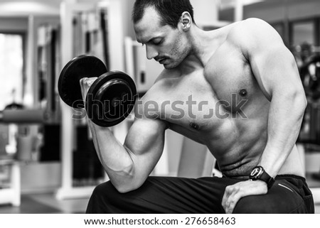 Muscular power athletic male bodybuilder sitting and training his biceps with dumbbells in fitness center. Black and white photo