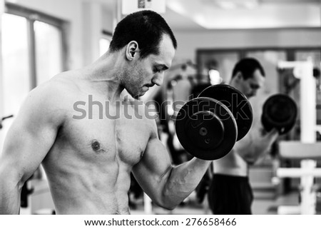Muscular power athletic male bodybuilder sitting and training his biceps with dumbbells in fitness center. Black and white photo