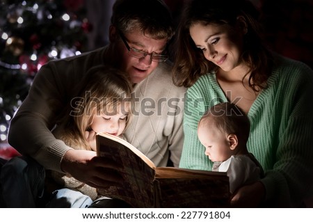 Family on Christmas Eve under the Christmas tree by the fireplace reading a book to their children with tales