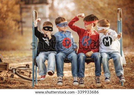 Four boys in superhero costume sitting on an old metal bed under a tree in the shade and boast super strength