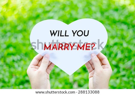 Hand holding white heart paper with will you marry me text on blur green grass background