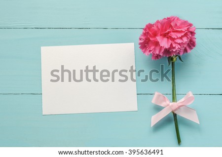 Blank white greeting card with pink Carnation flower and ribbon bow on blue wooden background