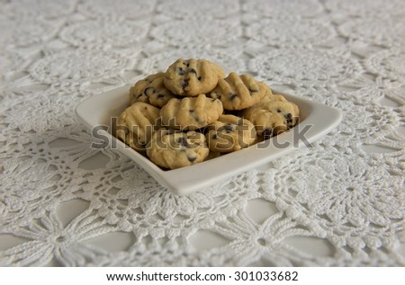 Chocolate chips cookies stacked up on a white plate.  Side view of the cookies displayed on a table covered by a white lace table cloth.