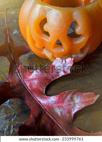 Halloween pumpkin wax candle with Red autumn leaf on a dirty, damaged, stained with wax, wood table.