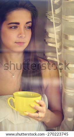 Beautiful young woman drinking her steaming coffee in front of the window and watching the city / Color effects, a soft film noise and a custom white balance is added to reach the Vintage look