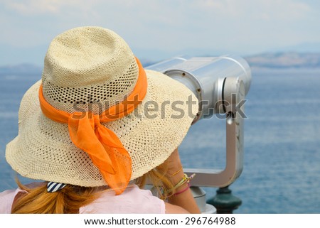 Young girl looking thru public binoculars at the seaside wearing straw hat and pink dress