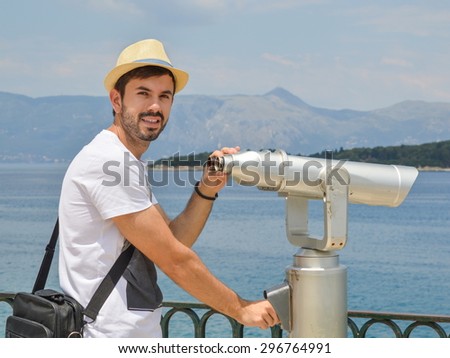 Young bearded man holding public binoculars at the seaside wearing straw on a sunny day