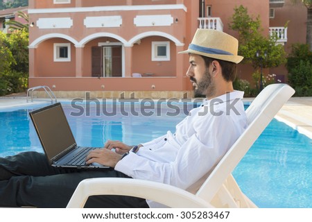 Young bussines man working on his lap top by the pool while on vacation  wearing straw hat