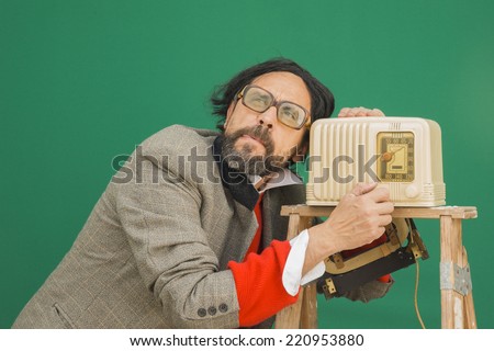 An untidy bizarre man, wearing big patched glasses and a toupee, listening to a radio station on an antique bakelite tube radio on a ladder, over green background
