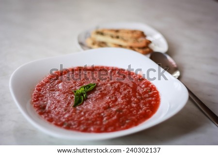 Cold tomato raspberry soup gazpacho decorated with green mint leaf in a white plate with crispy breads on the background