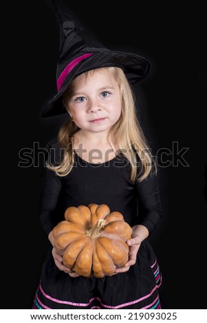 Halloween witch holding a pumpkin - portrait of little girl in black hat and black clothing on black background