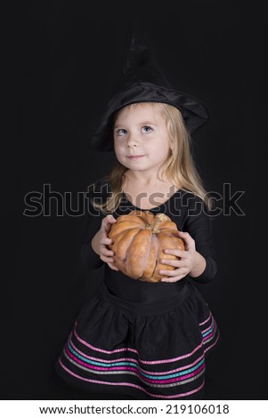 Portrait of little girl in black hat and black clothing with pumpkin on black background