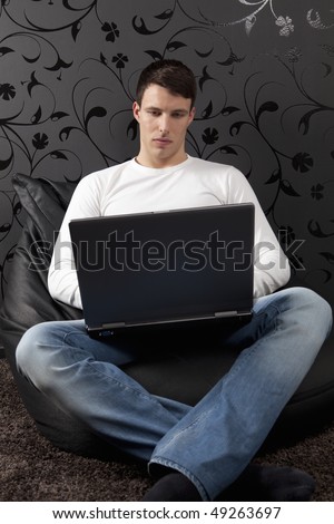 Handsome young man sitting on the carpet with the laptop, working. Black paper wall with ornaments behind.