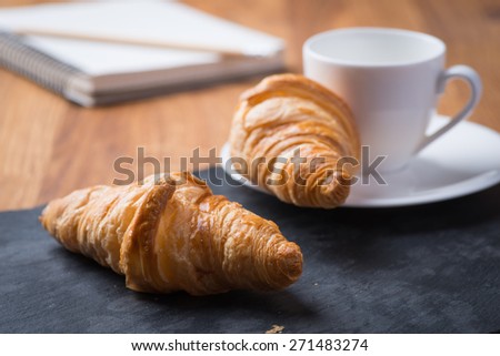 Two tasty and crusty french croissants with white cup of coffee and notebook on a wooden table