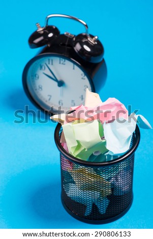 Small black trash bin with crumpled color paper inside and alarm clock over the blue background