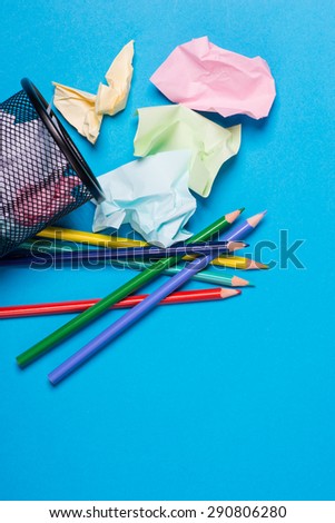 Small black trash bin with color pencils and crumpled color paper inside over the blue background