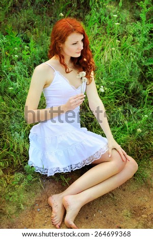 Cute redhead woman in white dress sitting on a grass in park with dandelions in her hands.