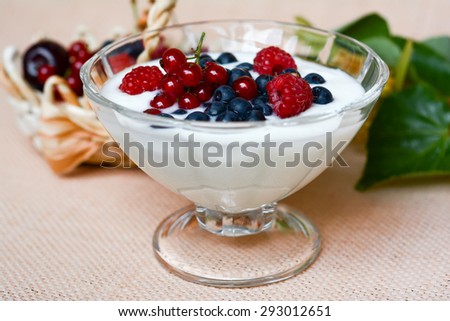 Yogurt with berries in glass bowl on sackcloth, beige background and basket with fresh berries and green leaf