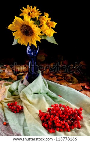 Sunflowers in a vase. The vase is on maple leaves. On the cloth are rowan berries. Autumn Still Life.