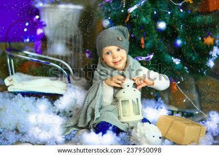 the girl in lilac jeans, in a knitted cap and with a knitted scarf sits on a sledge near a Christmas fir-tree with a toy and it is snowing