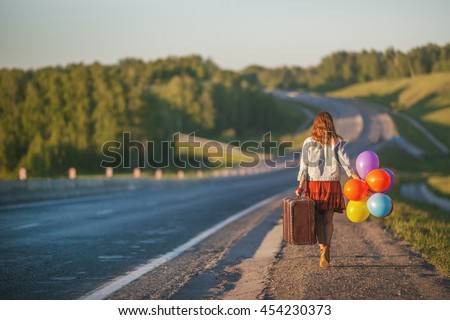 Pretty girl walking with big suitcase and colored balloons on the road. Sunlight, sunset, wildflowers, spring. Enjoying freedom and travel