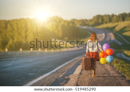 Pretty girl walking with big suitcase and colored balloons on field road. Sunlight, sunset, wildflowers, spring. Enjoying freedom and travel