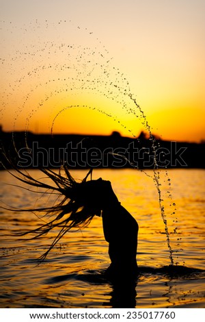 Silhouette of young fit woman posing standing in river playing with hair splashing water on sunset