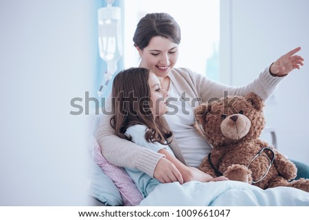 Older sister taking care of a sick child lying in a hospital bed with teddy bear