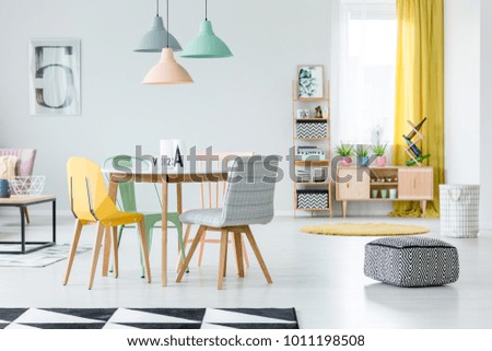 Patterned, black and white pouf in pastel dining room interior with colorful chairs standing at a wooden table